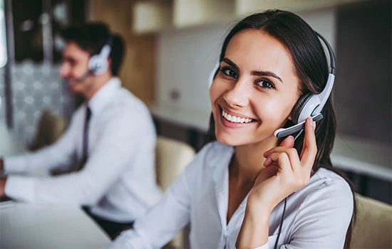 Young woman wearing headset smiling while talking on phone at office desk with male colleague working in background