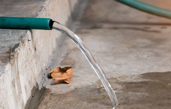 Water flowing from a green garden hose into a concrete basin with a brown autumn leaf floating inside
