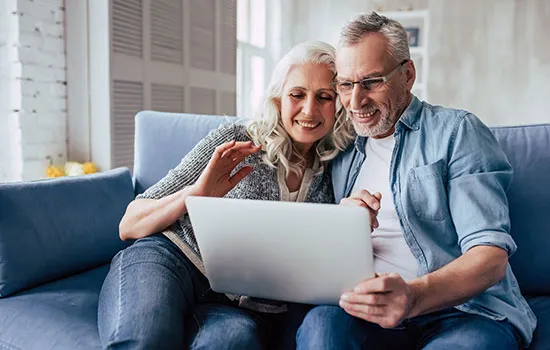 Mature couple smiling while using a laptop together on a blue couch in a bright living room