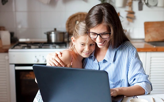 Adult woman and young girl looking at laptop screen together in kitchen, both smiling while using computer