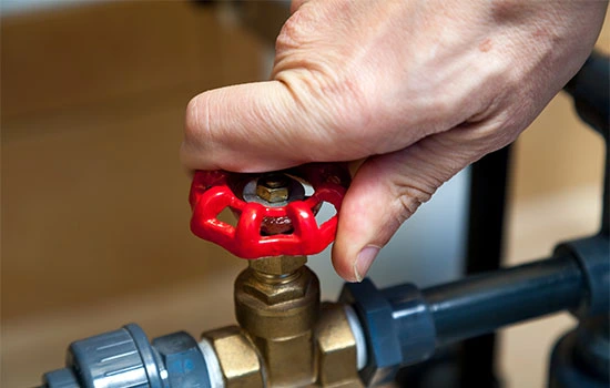 Hand turning a red ball valve on a brass pipe fitting connected to blue plumbing tubing
