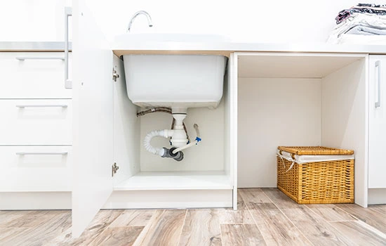 Open white cabinet under sink with plumbing pipes visible, wicker storage basket on right side, wooden floor, white walls and cabinetry