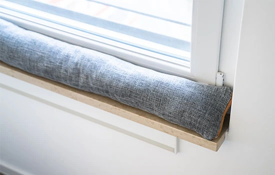 Gray fabric draft stopper installed at base of window with striped Roman shade, sitting on wooden sill to block drafts and reduce energy loss