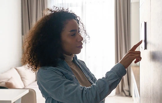 Woman with curly hair wearing a denim shirt pointing at a wall-mounted thermostat in a bright, modern bedroom with white bedding and curtains.